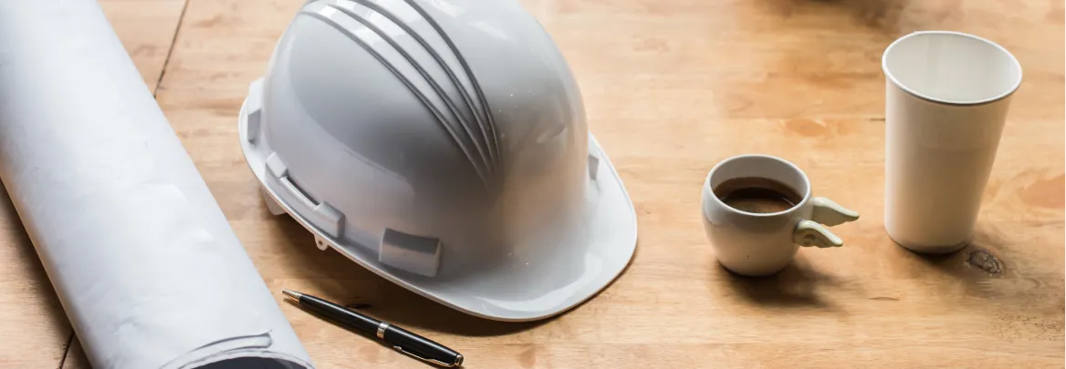 Photographie qui met en scène une pause café dans l'univers de la construction Alternative Bois Concept, un casque étant posé sur une table à côté d'un café et de plans de chantiers.